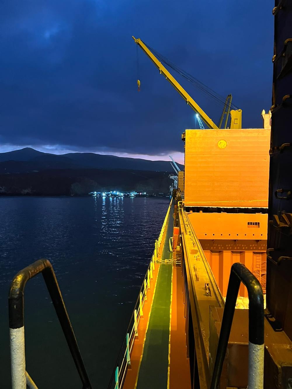 Cubierta de un buque de carga general de noche, con grúa de a bordo iluminada y la bahía al fondo